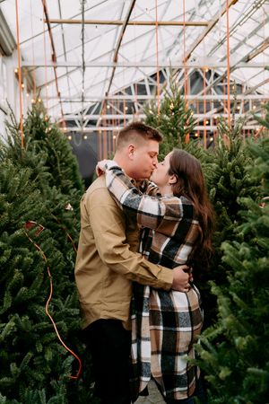A couple shares a romantic moment among rows of Christmas trees in a greenhouse.