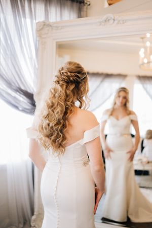 Bride in fitted white off-shoulder gown with curled hair looking in mirror during wedding preparations.