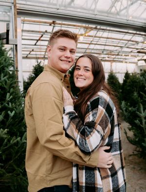 Romantic couple embraces in a greenhouse filled with evergreen trees during a winter photo session.