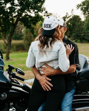 A couple embraces near a motorcycle, with one wearing a denim jacket and 'BRIDE' baseball cap in an outdoor setting.