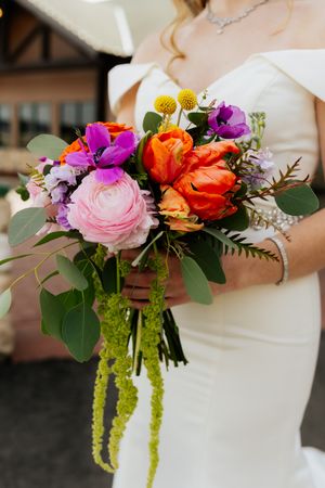 A vibrant bridal bouquet featuring pink, orange, and purple flowers with cascading greenery and yellow craspedia.