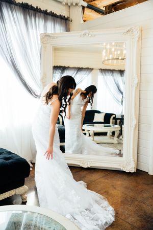 Woman in white wedding dress looking in ornate white framed mirror with elegant curtains and vintage decor.