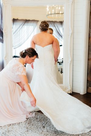 A white wedding dress is being adjusted as the train flows across a gray carpet in an elegant dressing room.