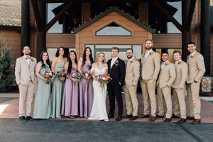 A bridal party poses outdoors wearing soft pastel and beige colors in front of a rustic wooden entrance.