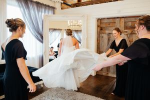 Wedding party attendants in black dresses helping to fluff and arrange a flowing white wedding gown in a rustic venue.