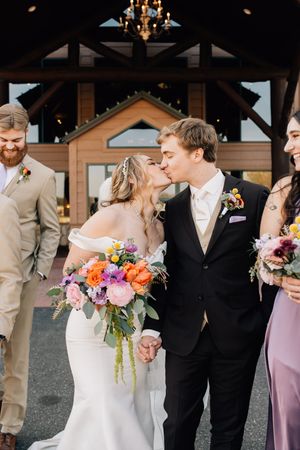 Joyful wedding party gathers outside rustic venue with bride holding vibrant orange and pink floral bouquet.