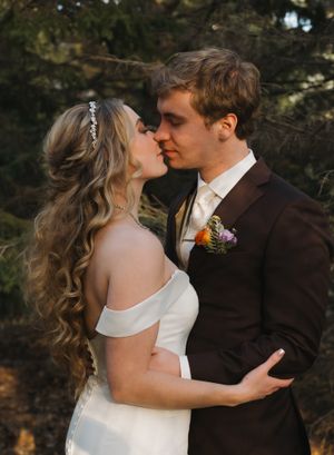 A romantic wedding portrait of a couple sharing a kiss outdoors with the bride in white and groom in brown suit.