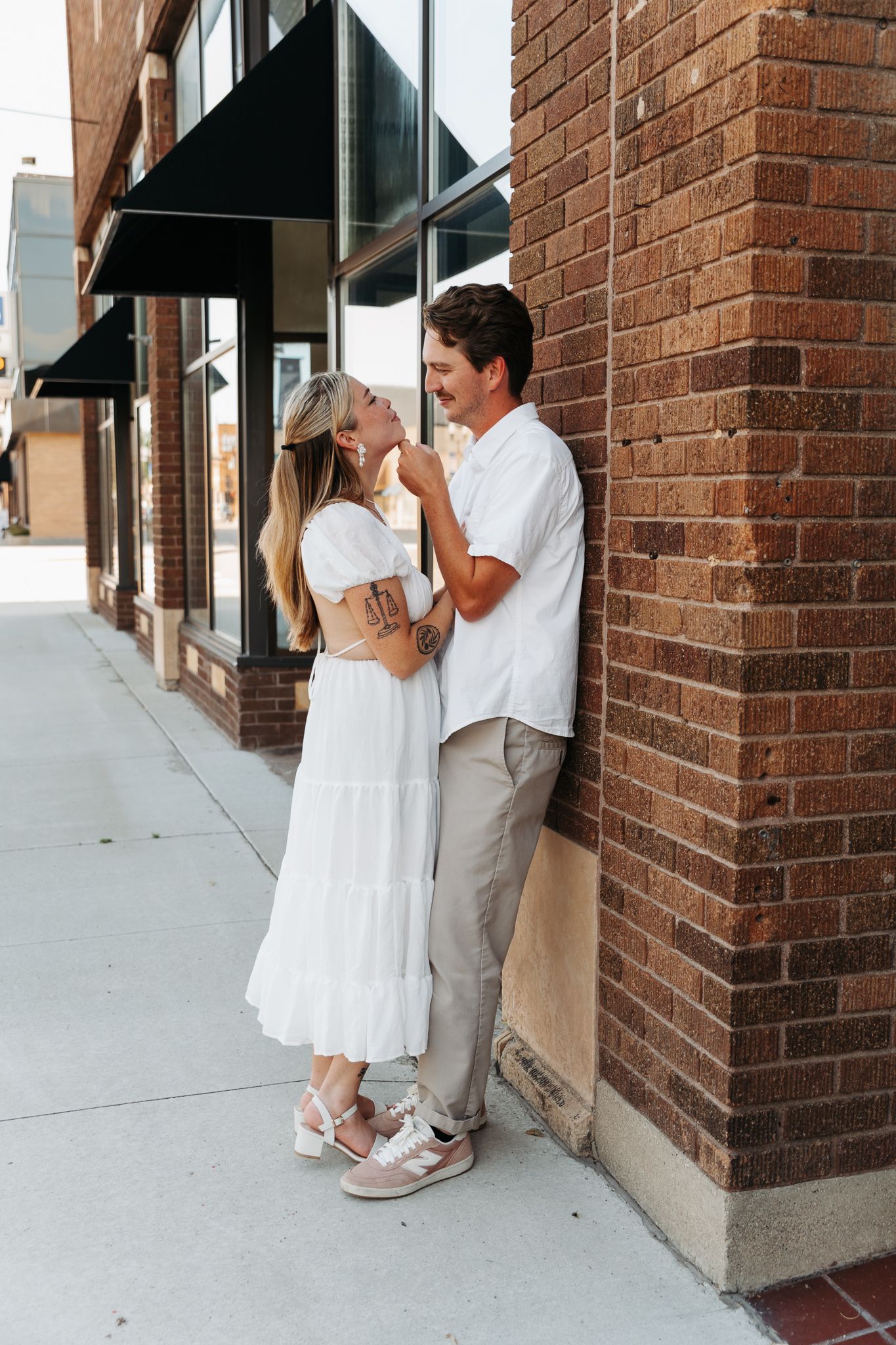 Couple in white outfits share playful moments against a brick building wall during a photo session.