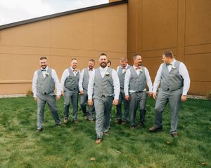 Groomsmen in matching light gray vests and slacks stand together on grass in front of a tan wall.