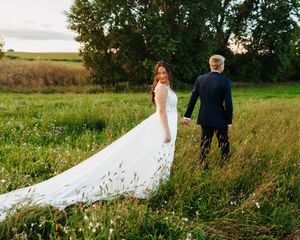 A couple walks through a scenic field with the bride's white dress flowing behind them in the golden sunlight.