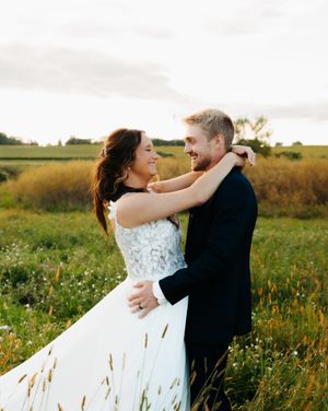 Romantic couple embraces in a sunlit meadow with wildflowers as her white wedding dress flows in the breeze.