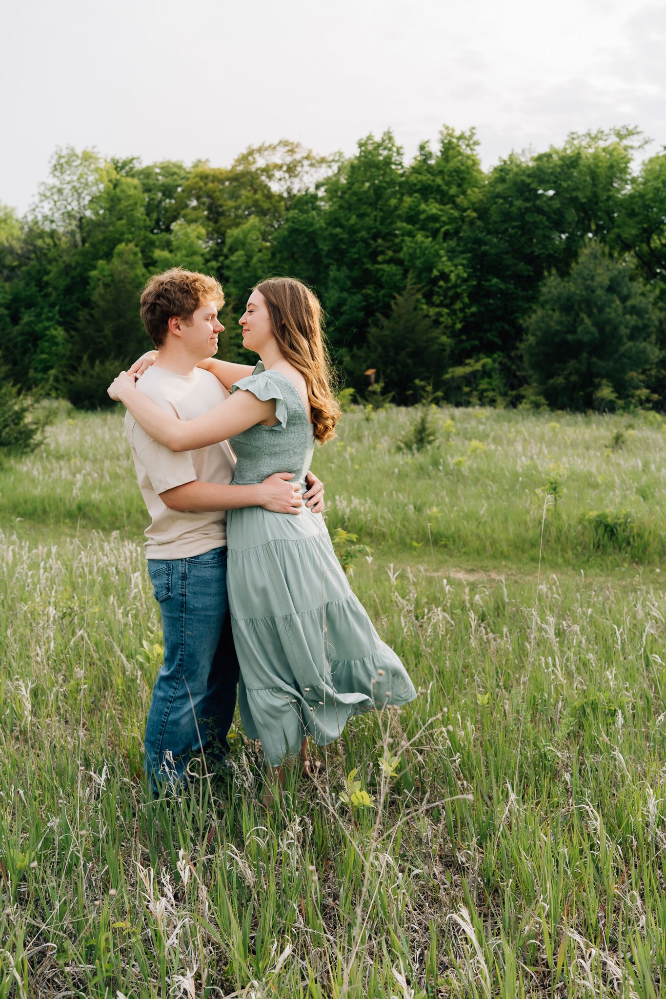 A romantic couple embraces while dancing together in a sunlit meadow filled with tall grass and wildflowers.