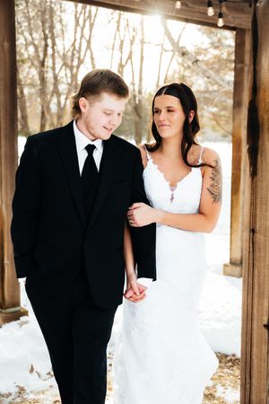Couple in wedding attire share an intimate moment under a wooden archway in the winter snow.