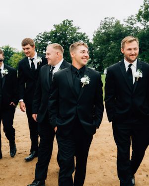 Group of groomsmen in black suits standing together on sandy ground with trees in background.