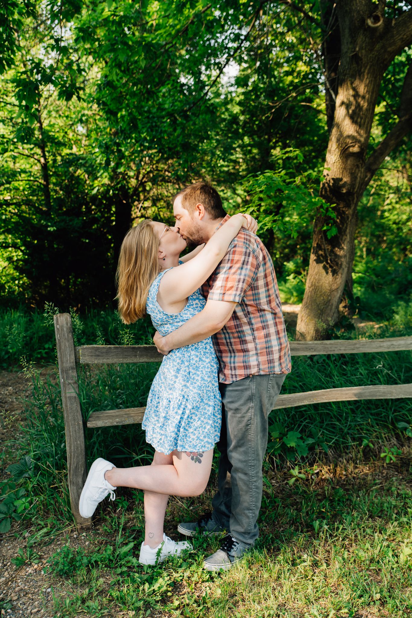A romantic couple sits together on a wooden fence surrounded by lush green trees and foliage in a park setting.