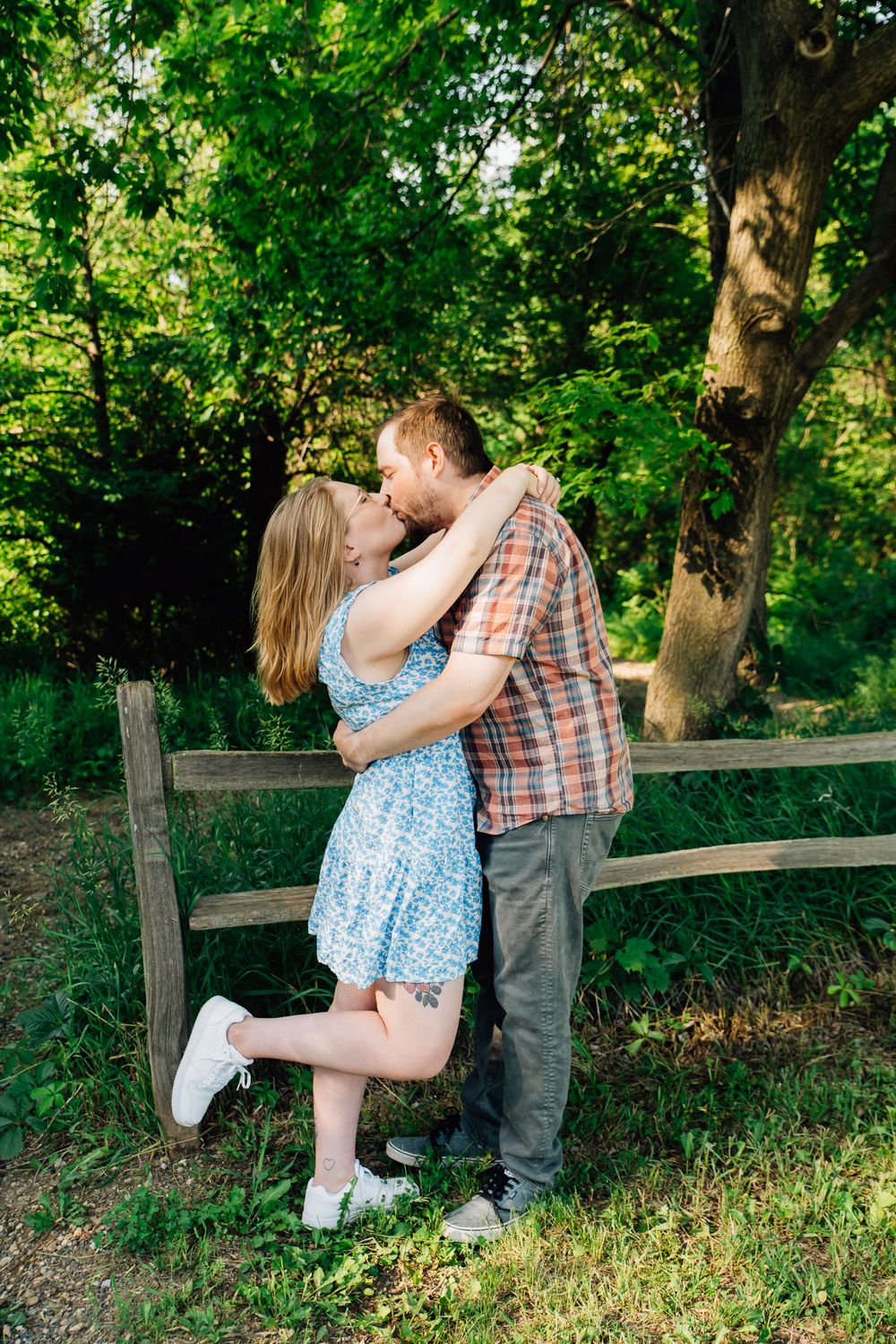 A romantic couple sits together on a wooden fence surrounded by lush green trees and foliage in a park setting.