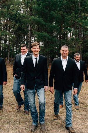 A group of men in mixed formal and casual attire walk through a wooded area.