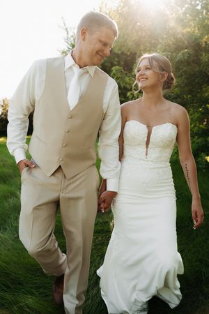 Newlyweds in elegant wedding attire walking together on grass during golden hour.