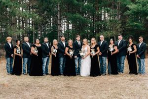 Wedding party in black and navy formal attire poses together in front of pine trees at an outdoor venue.
