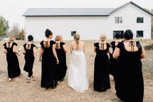 A bride in white and bridesmaids in black dresses walk together toward a rustic barn on a rural property.