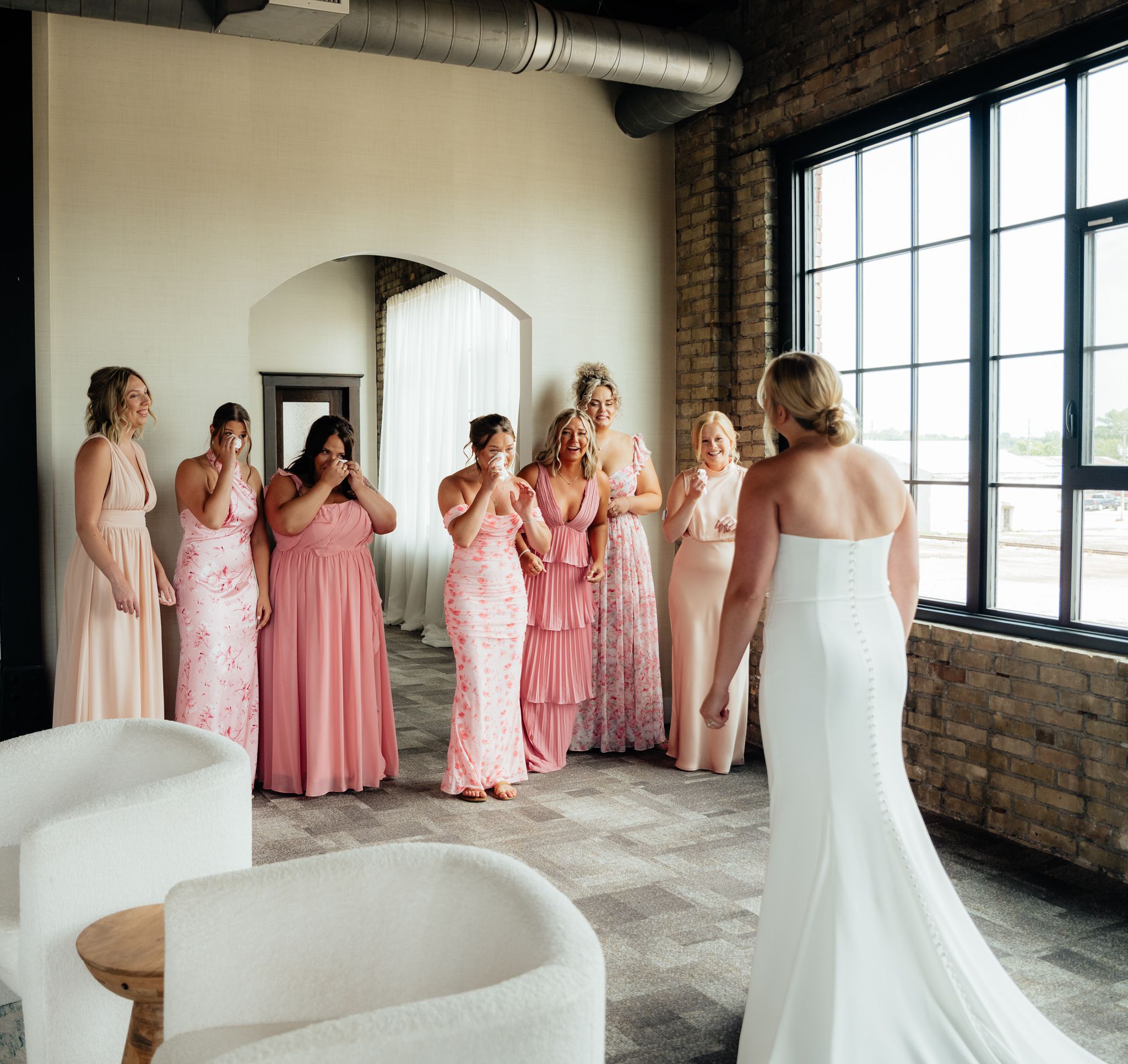 Bridesmaids in pink dresses gather around a bride in a white gown in an industrial-style venue.