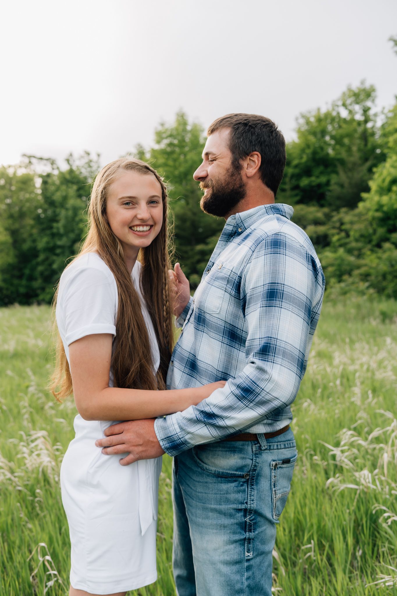 Romantic couple embracing in a sunny meadow with tall grass and trees in the background.