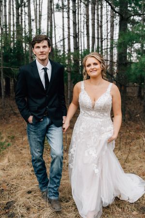 A couple in wedding attire poses in a pine forest, with the groom wearing jeans and the bride in a white lace dress.