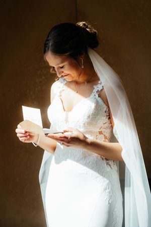 A backlit bride in a lace wedding gown reads a letter while wearing her veil during a quiet moment.