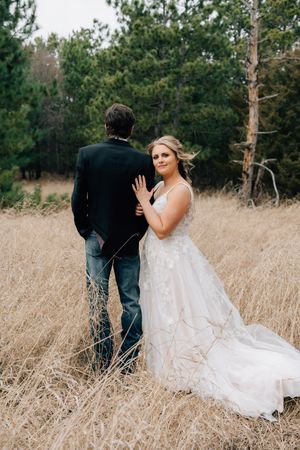A couple in wedding attire poses in a field of tall grass with evergreen trees in the background.