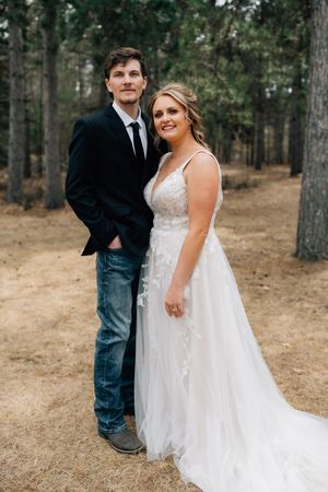 Wedding couple poses together in a forest setting with the bride wearing a lace white gown and groom in suit jacket and jeans.