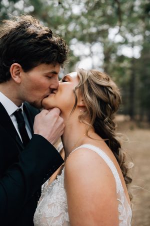 A couple shares a tender kiss in a romantic outdoor wedding setting with soft natural light filtering through trees.