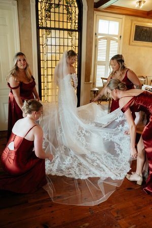 Bridesmaids in burgundy dresses help arrange a delicate lace wedding veil on hardwood floors.