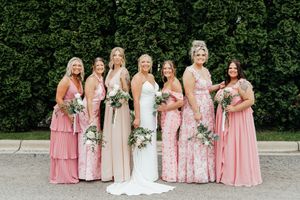 A bridal party poses in pink and neutral bridesmaid dresses against a lush green hedge backdrop.