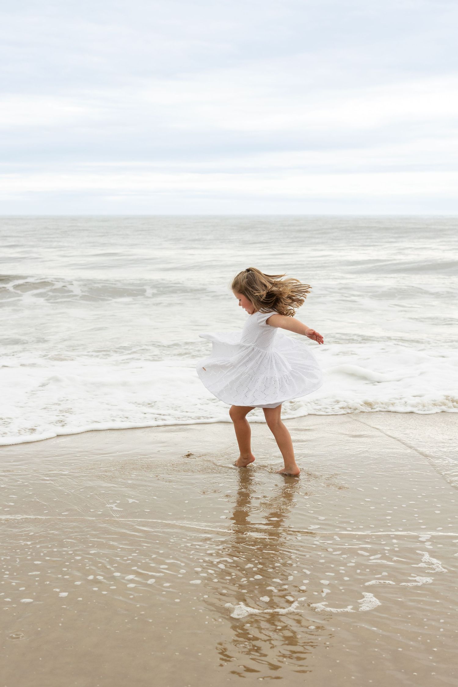 Child in white dress twirling and dancing on a beach shore as waves crash in the background.