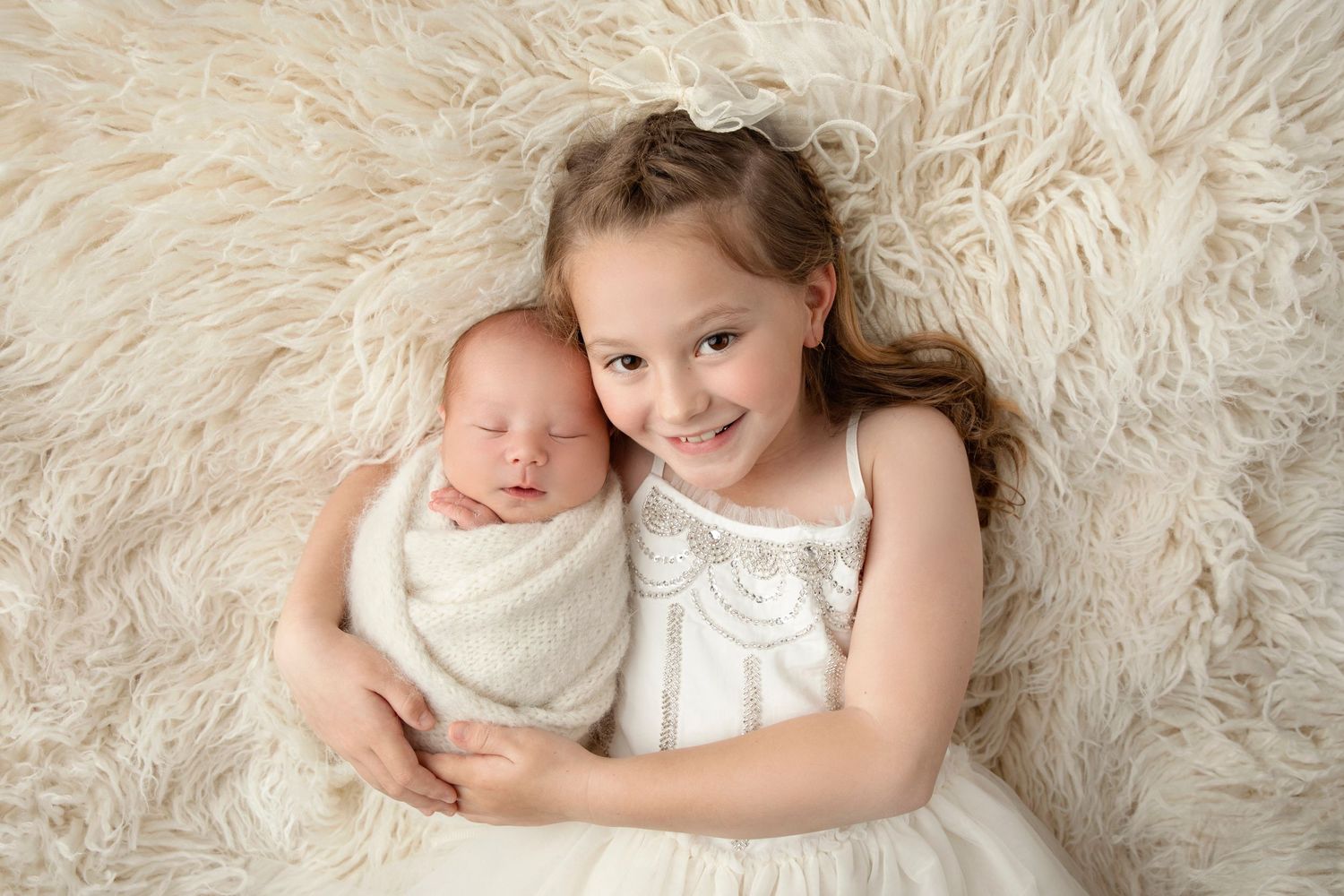 Sweet siblings portrait on fluffy white rug with newborn and older sister.