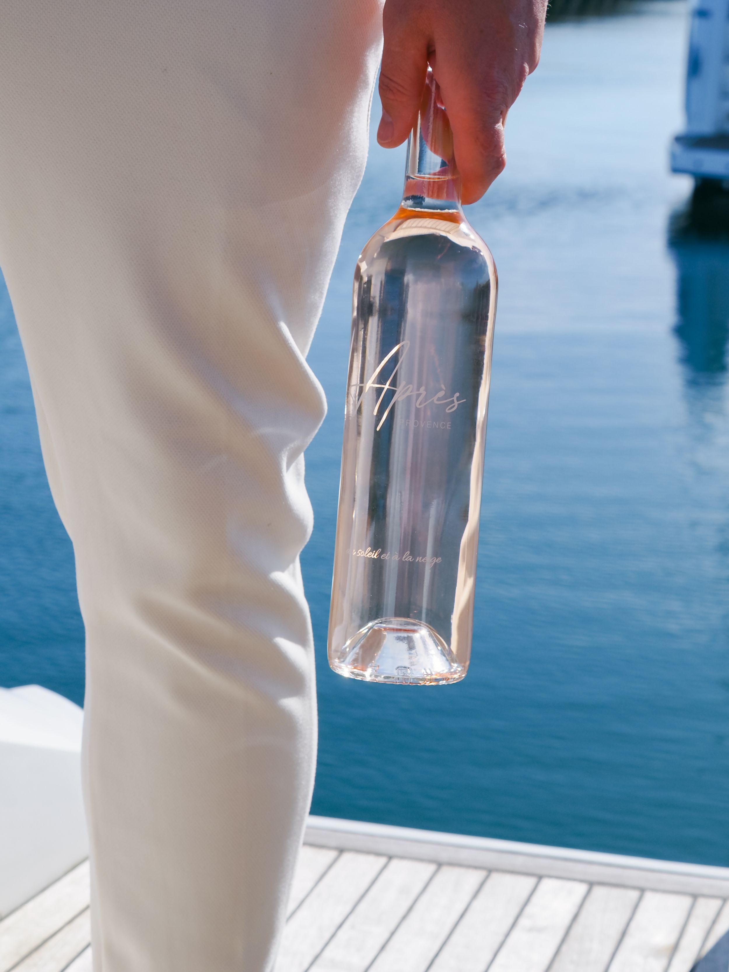 Person in white pants holding a glass bottle while standing on a wooden dock by blue water.