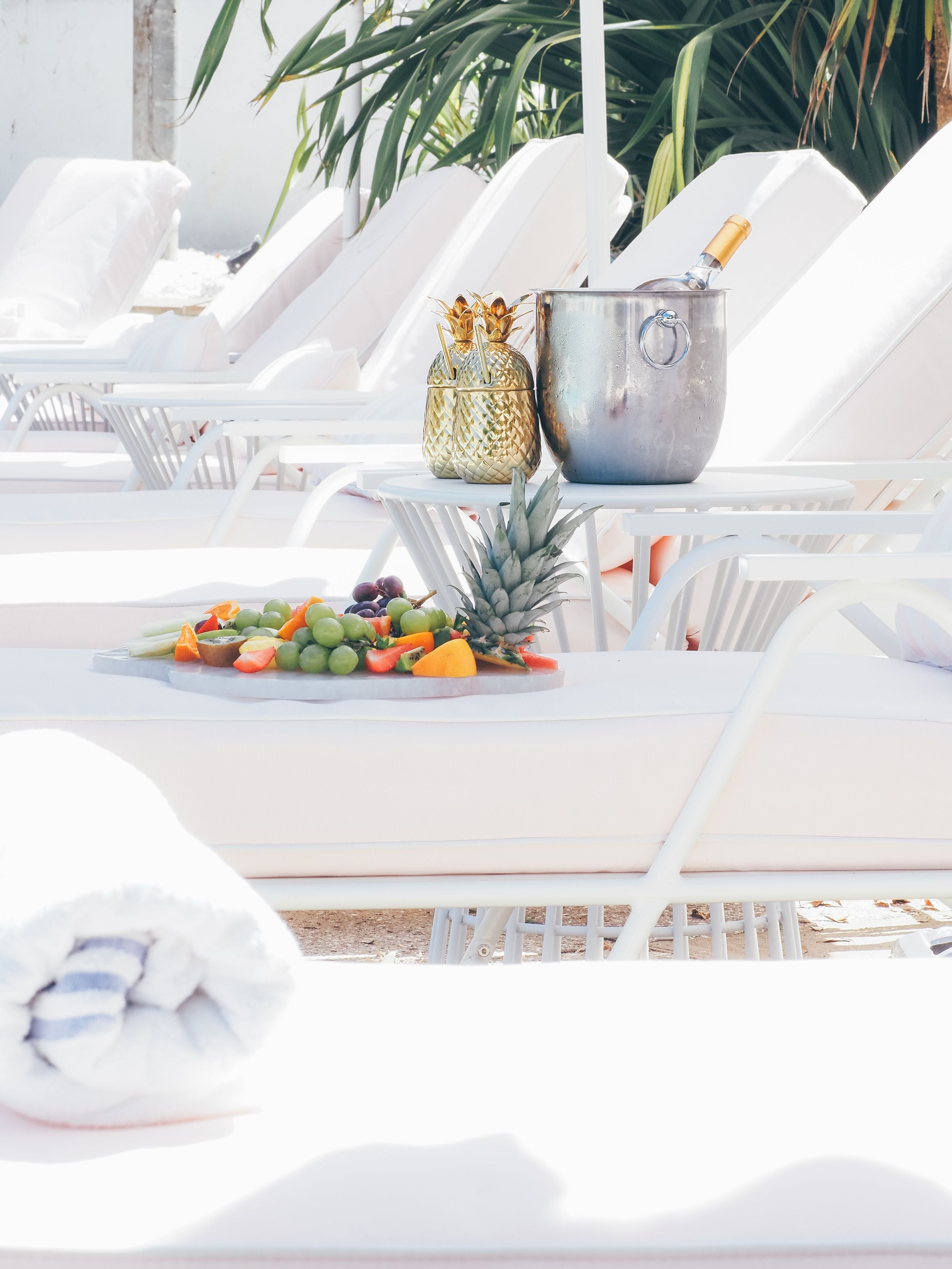 Luxury poolside setup with white loungers, tropical fruit platter, and decorative gold pineapple container near palm trees.