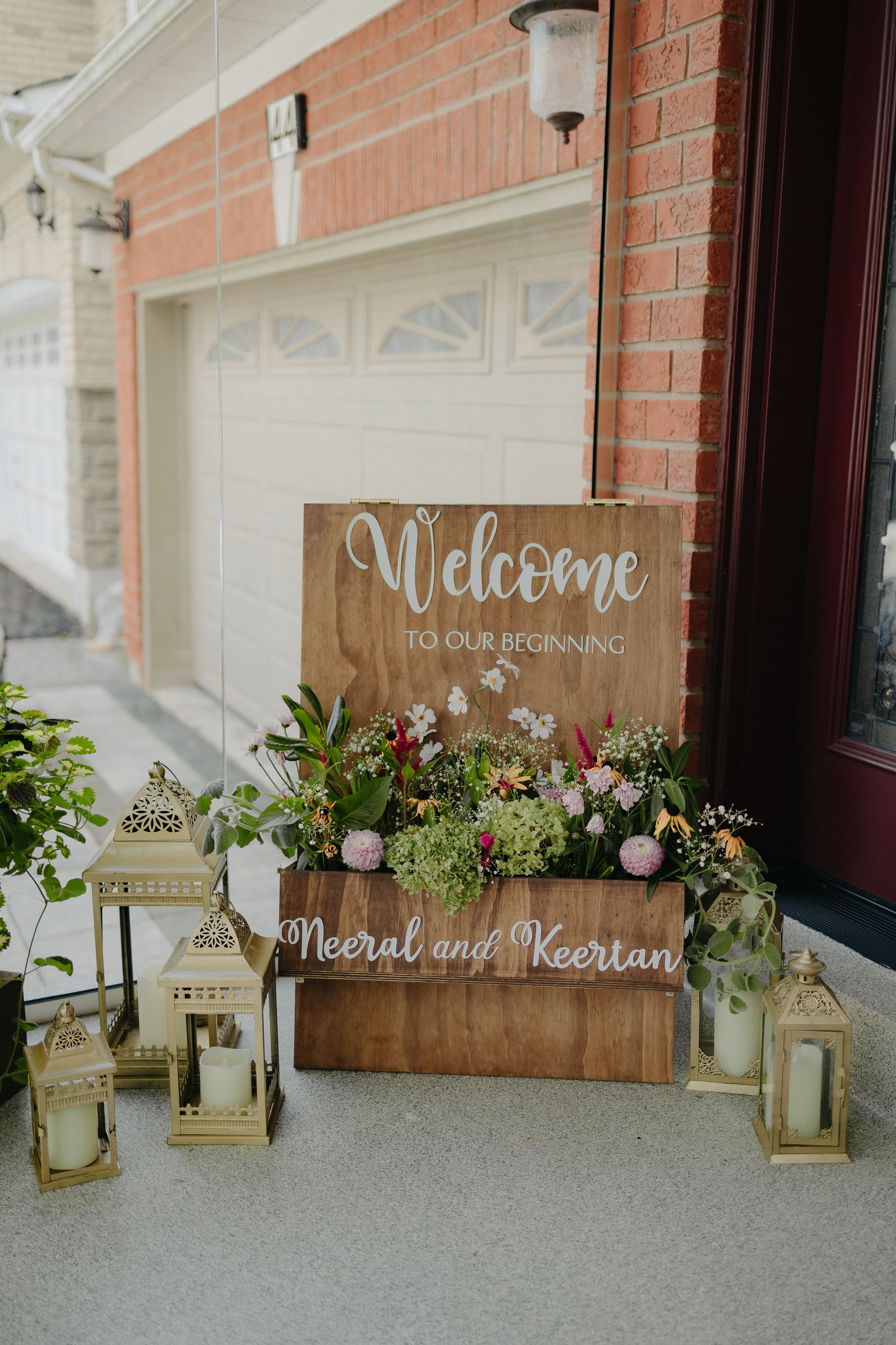 A rustic wooden welcome sign with lanterns and floral decorations at a wedding entrance.