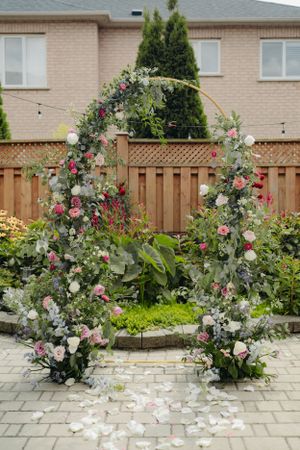 A floral arch decorated with pink roses and white flowers stands in a backyard garden with a wooden fence.