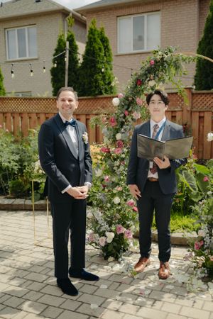 An intimate outdoor wedding ceremony setup with a floral arch and brick house backdrop.