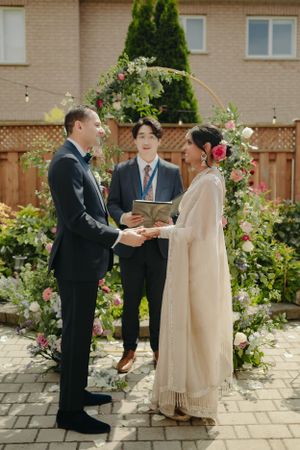 An intimate wedding ceremony taking place under a floral arch in a backyard setting.