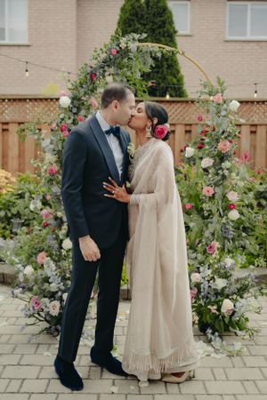 A romantic moment under a floral arch decorated with pink and white roses in a backyard garden setting.