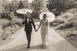 A couple walks hand in hand with parasols in a black and white garden photo.