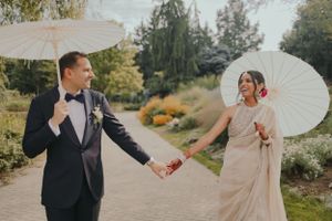 A playful couple holds hands while walking with parasols in a garden setting.