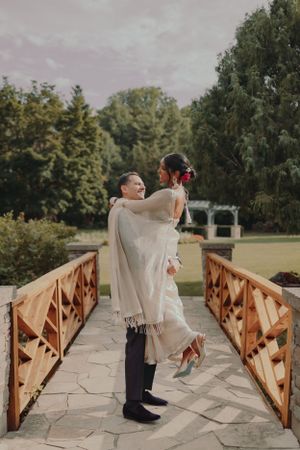Romantic wedding couple embraces on a wooden bridge surrounded by lush garden scenery.