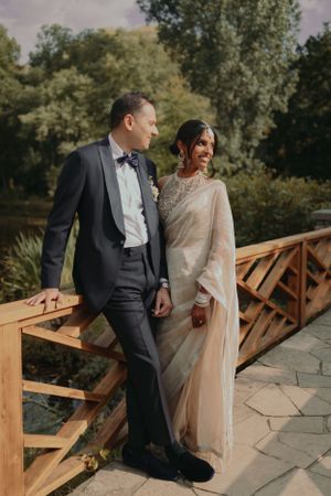 Elegant couple in wedding attire pose together on a wooden bridge in a garden setting.