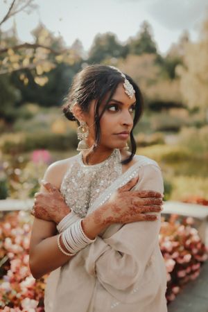 Bride in traditional white sari with henna adorned hands poses in garden setting.