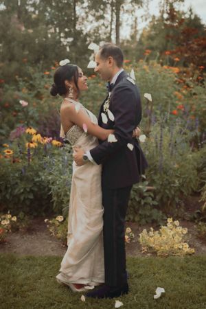 A romantic wedding moment in a garden with petals falling as the couple embraces in formal attire.