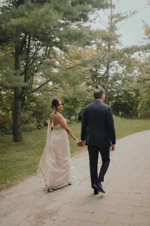 Newlyweds walking hand in hand down a tree-lined path in elegant wedding attire.