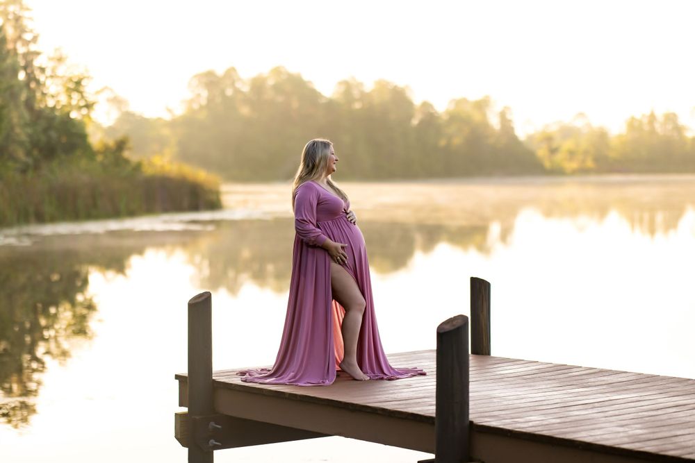 A figure in a flowing purple dress stands on a wooden dock at sunset overlooking a peaceful lake.